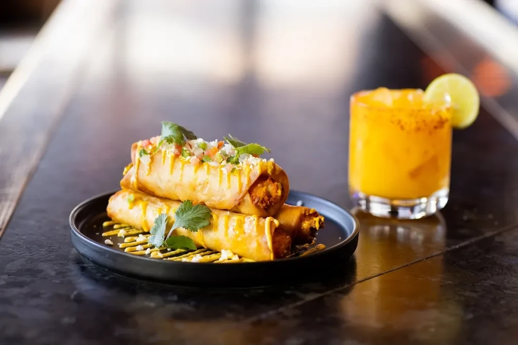 Close-up of three topped taquitos and an orange drink with lime.