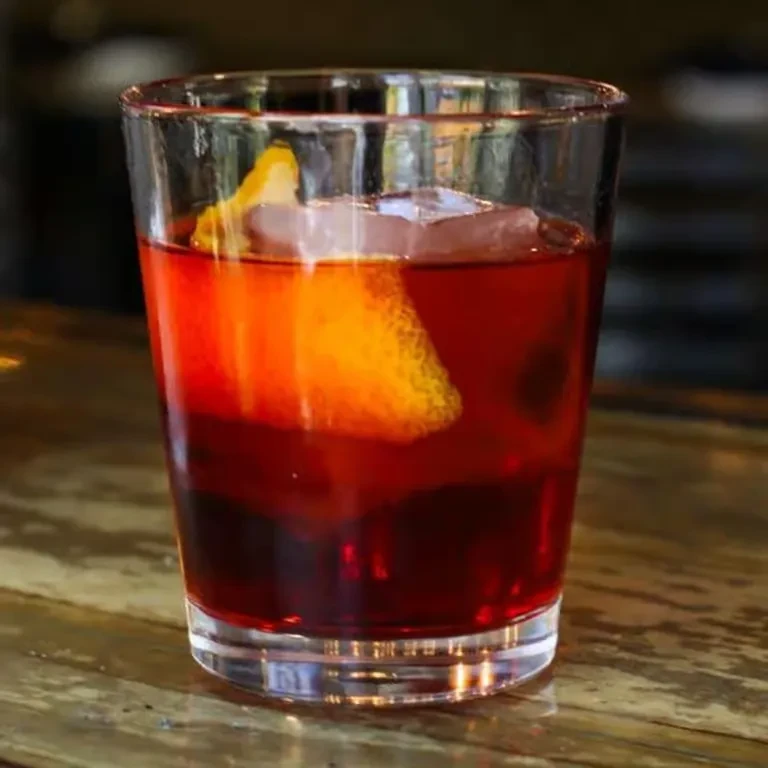 Red cocktail with ice and a citrus twist on a rustic wooden bar.
