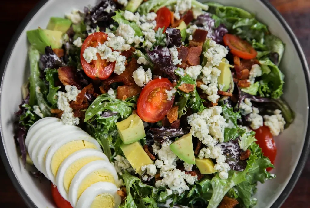 Overhead colorful salad with avocado, bacon, egg, and blue cheese.