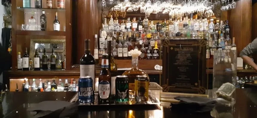 Dimly lit bar counter with extensive liquor selection and hanging glassware.