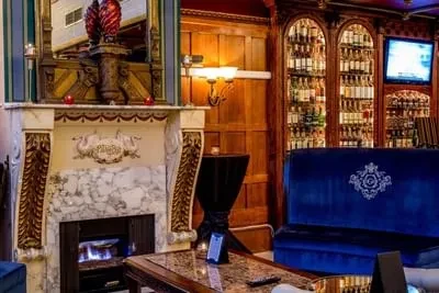 Grand fireplace, stocked bar, and blue velvet sofa in an upscale lounge.