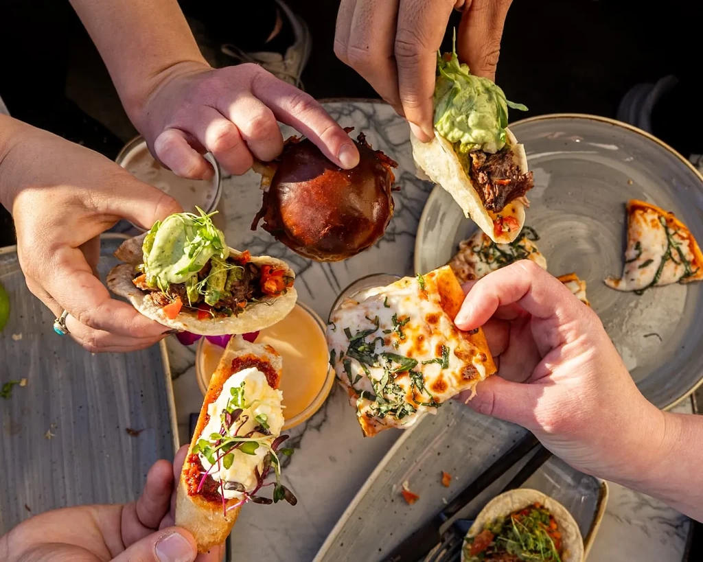 Overhead view: multiple hands sharing tacos, pizza, and bruschetta.