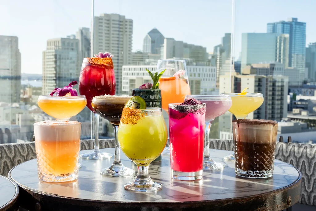 Nine diverse cocktails on a table overlooking a modern cityscape.