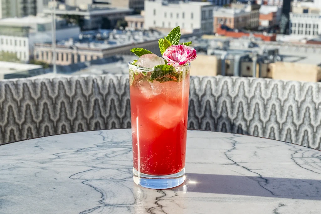 Reddish-pink cocktail with mint and flower on marble table, city view.