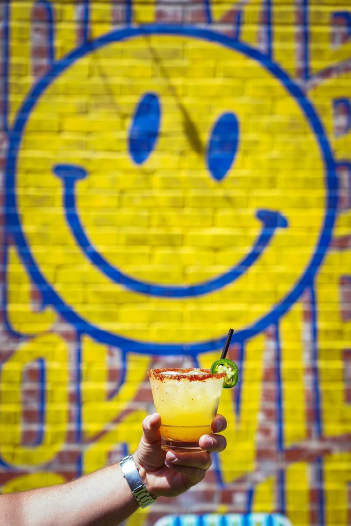 Hand holding yellow cocktail with jalapeño, spiced rim, by graffiti.
