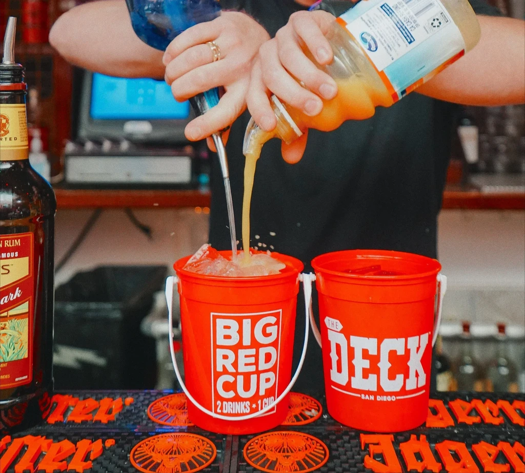 Hands pouring drinks from a shaker and bottle into a red cup.