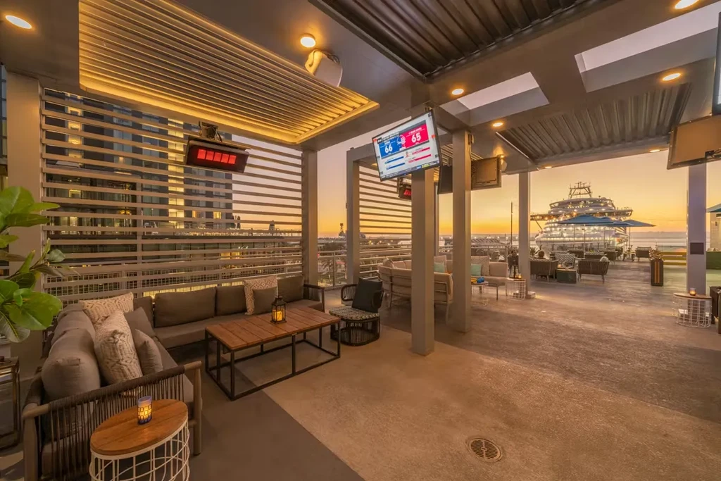 Outdoor lounge with heaters, TVs, city skyline, and cruise ship at sunset.