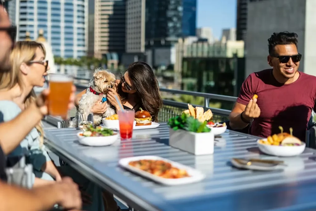 Friends dining on a rooftop patio overlooking a cityscape, with a dog.