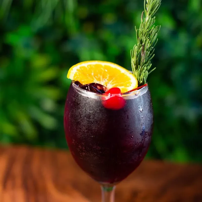 Cold red cocktail in wine glass with citrus slice, cherry, rosemary.