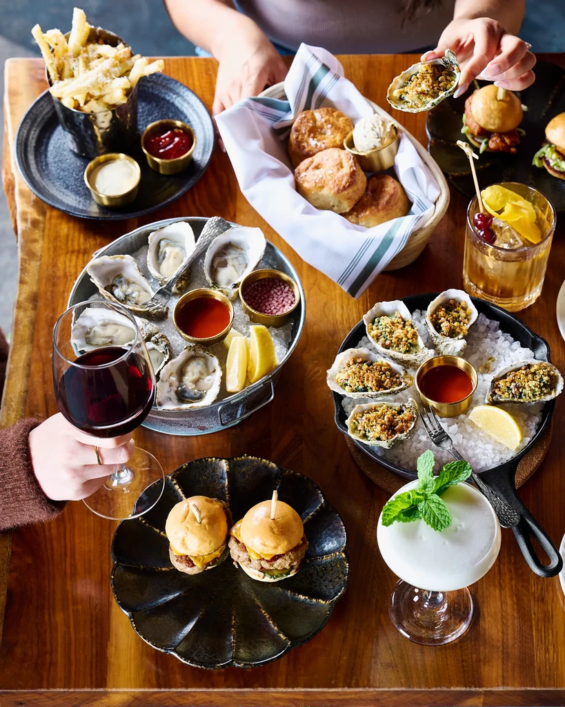 Top-down view: fries, oysters, sliders, biscuits, cocktails on wooden table.