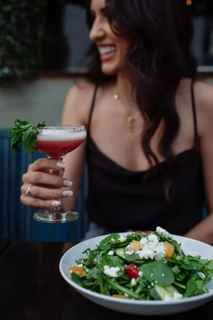 Hand holding pink cocktail and colorful salad; smiling woman behind.