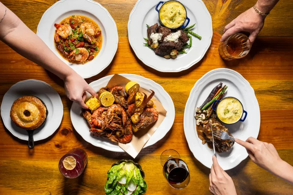 Hands eating various dishes and drinks at a wooden dining table.