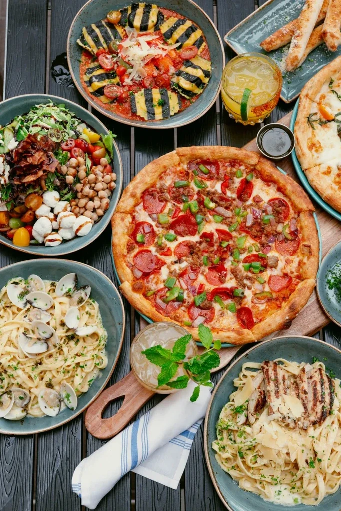 Overhead flat lay of Italian pizzas, pastas, salad, and cocktails.