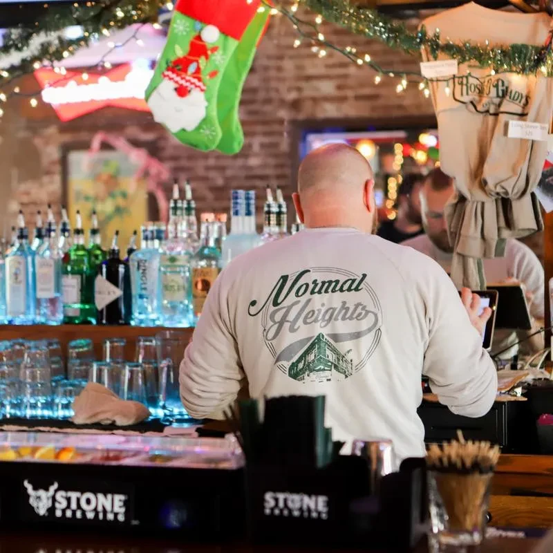 Bartender in "Normal Heights" shirt at blue-lit bar with Christmas decorations.