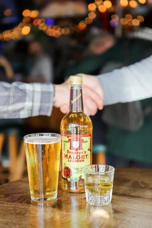 Malört bottle, beer, and shot glass on a wooden table, blurred background.