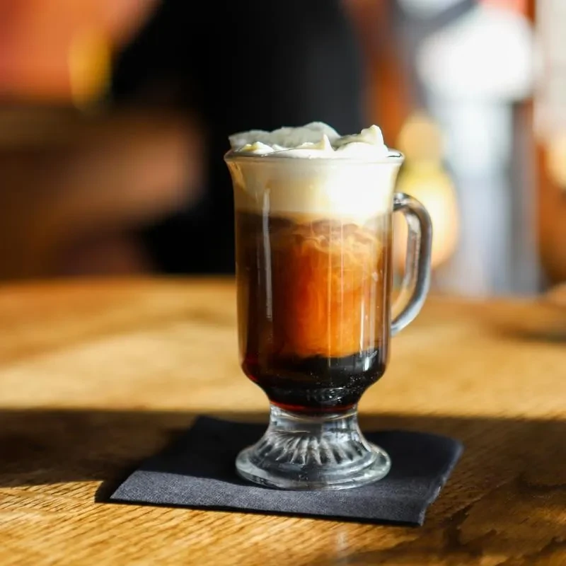 Layered drink with dark, amber, and whipped cream in a tall glass.