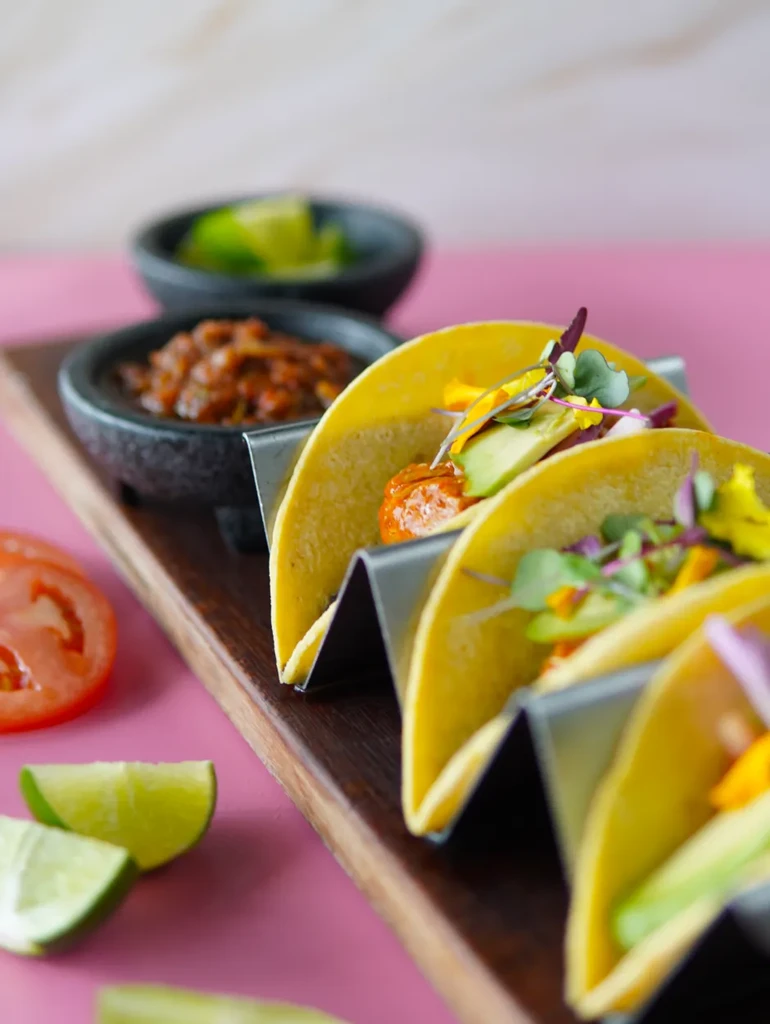 Vibrant tacos with avocado, protein, and microgreens on a pink surface.