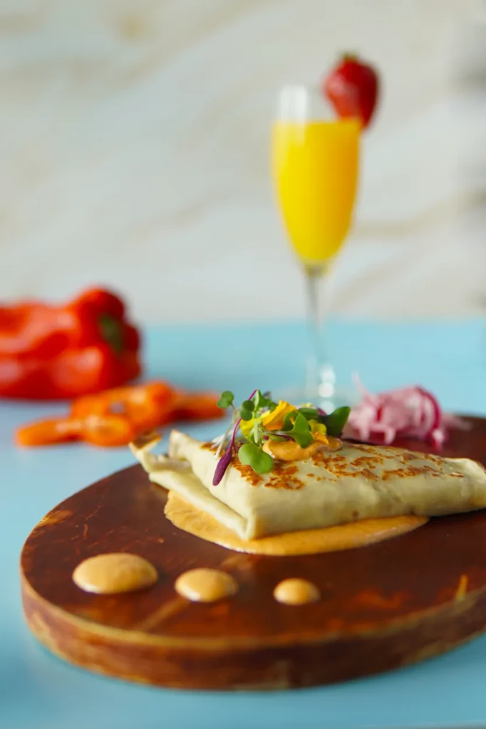 Savory crepe with orange sauce, microgreens, and edible flowers on a wooden board.