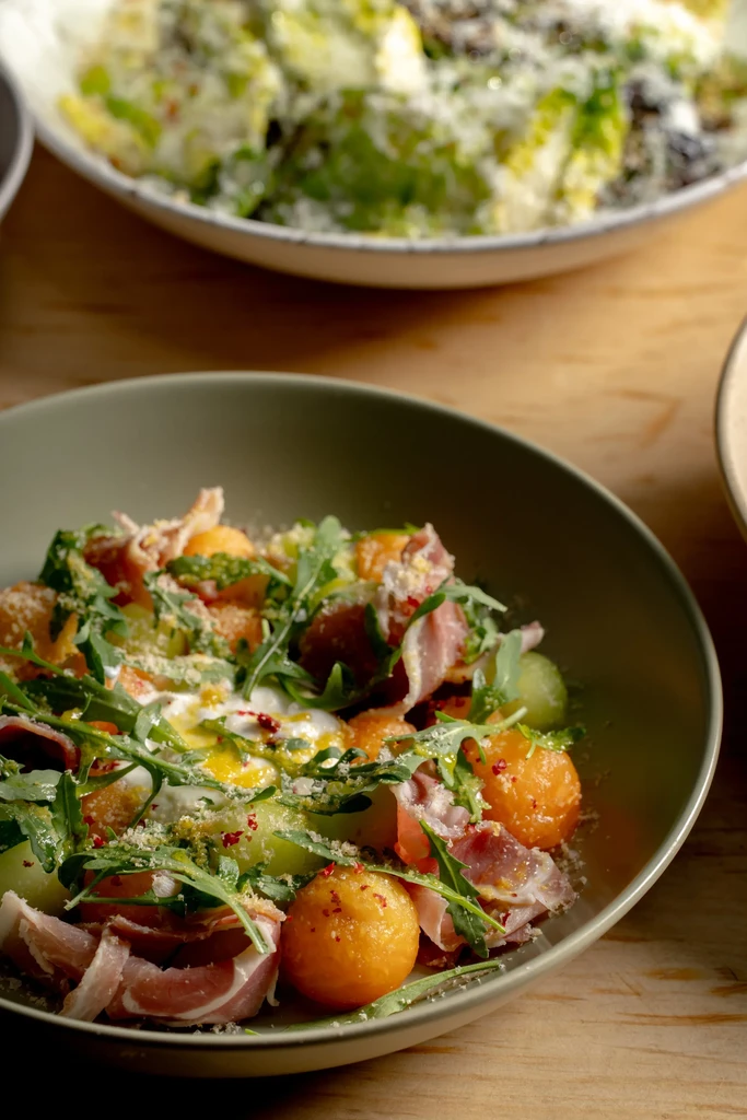 Close-up of arugula, prosciutto, melon salad; another out-of-focus salad.