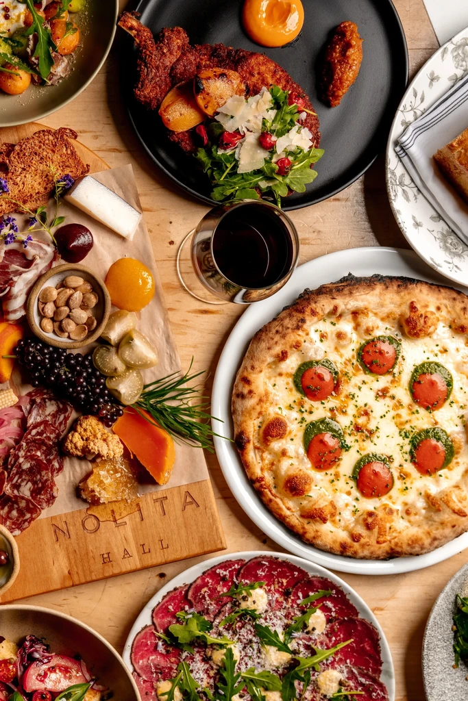 Overhead flat lay: pizza, charcuterie, and dishes on a 'NOLITA HALL' board.