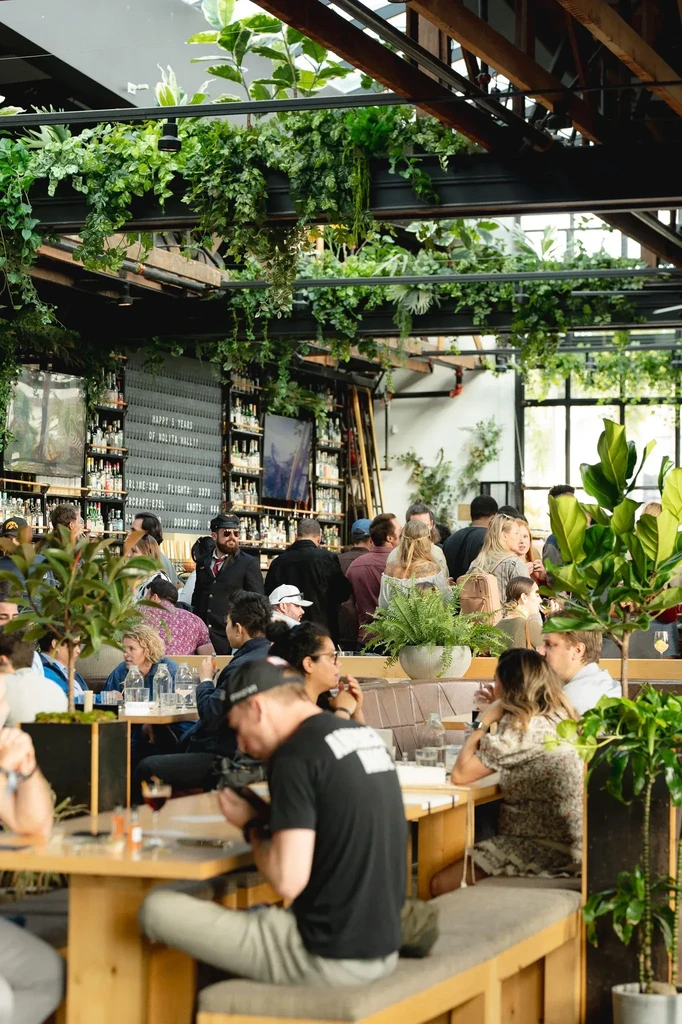 Lively industrial-chic restaurant interior with abundant green plants and patrons.