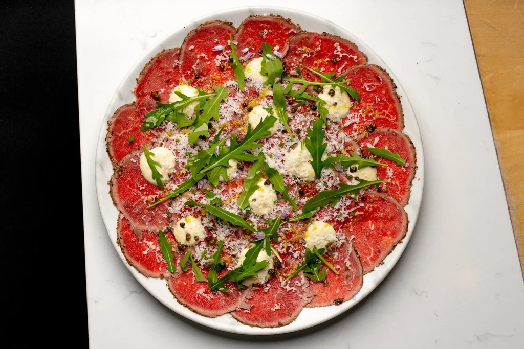 Overhead shot of beef carpaccio with Parmesan, arugula, sauce, and peppercorns.
