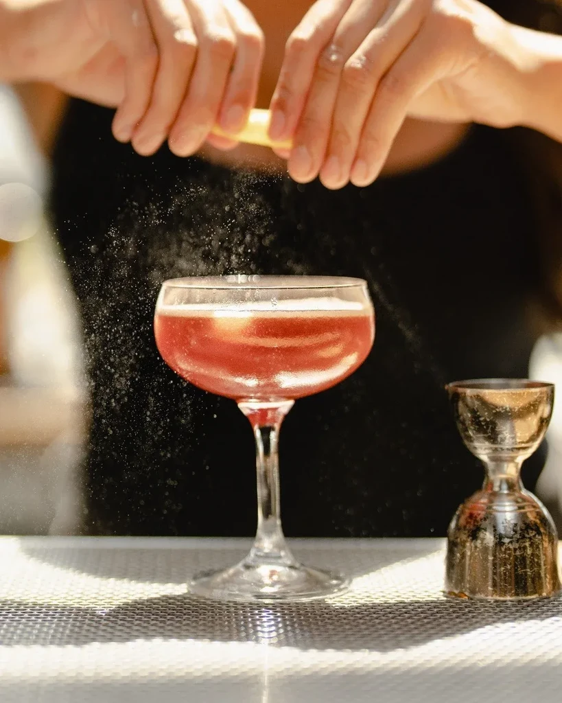 Hands squeeze citrus peel, misting zest over a pink cocktail.