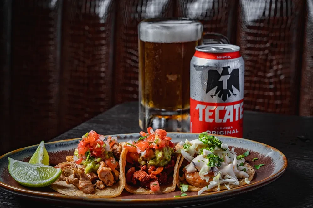 Three diverse tacos, a beer glass, and a Tecate can.