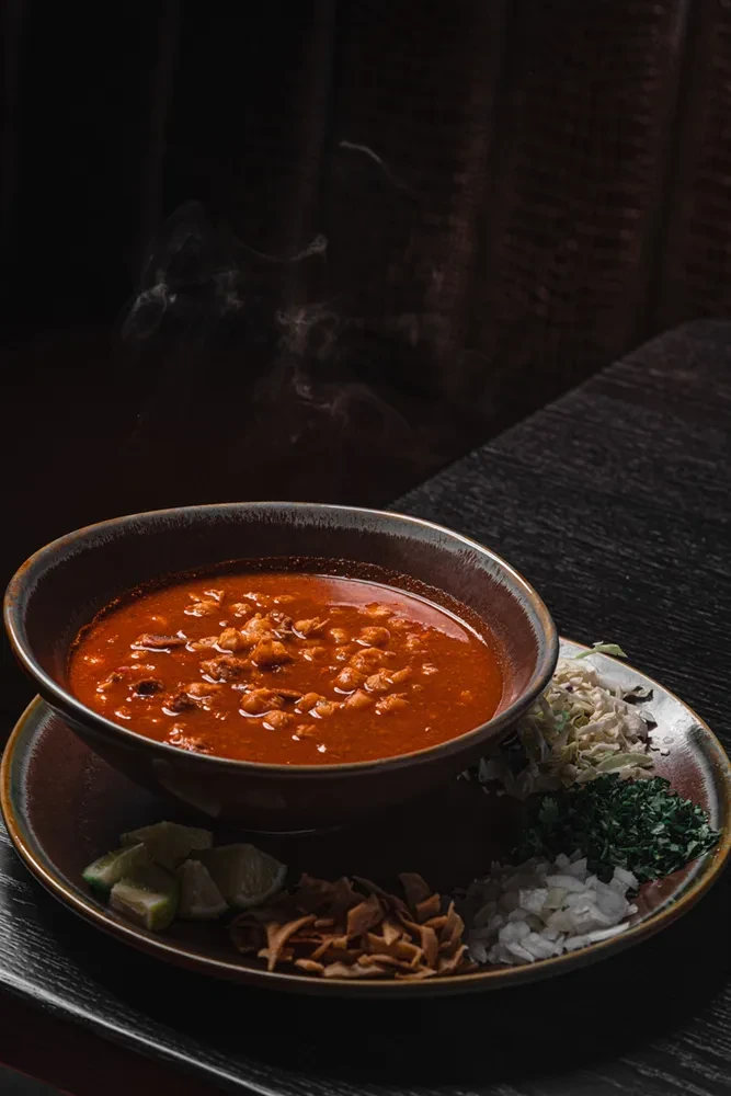 Steaming bowl of red pozole with garnishes on a dark rustic plate.