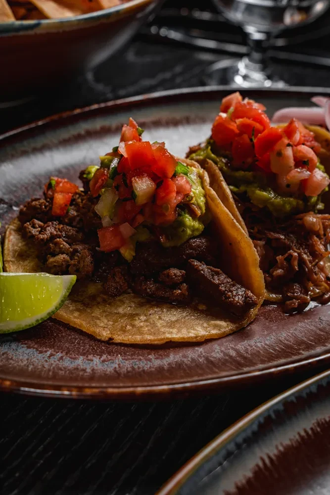 Two tacos filled with meat, guacamole, pico de gallo, and lime wedge.