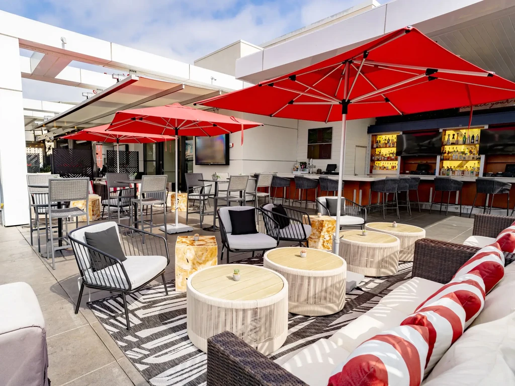 Outdoor modern restaurant patio with varied seating, red umbrellas, and visible bar.