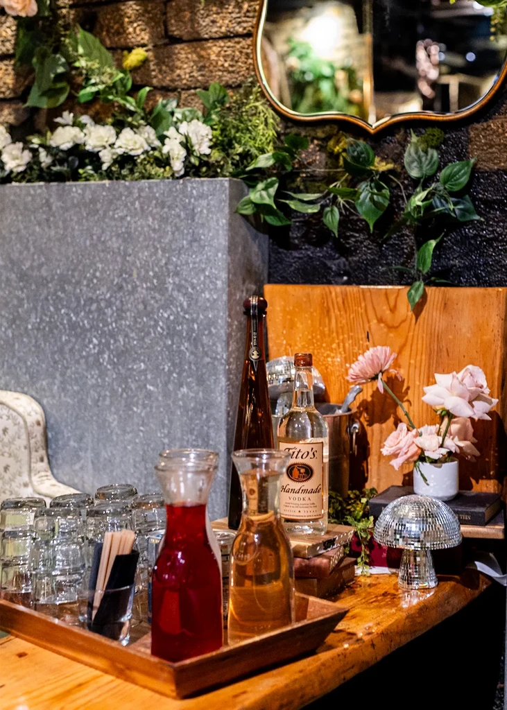 Rustic wooden bar with various bottles, drinks, flowers, and disco mushroom.