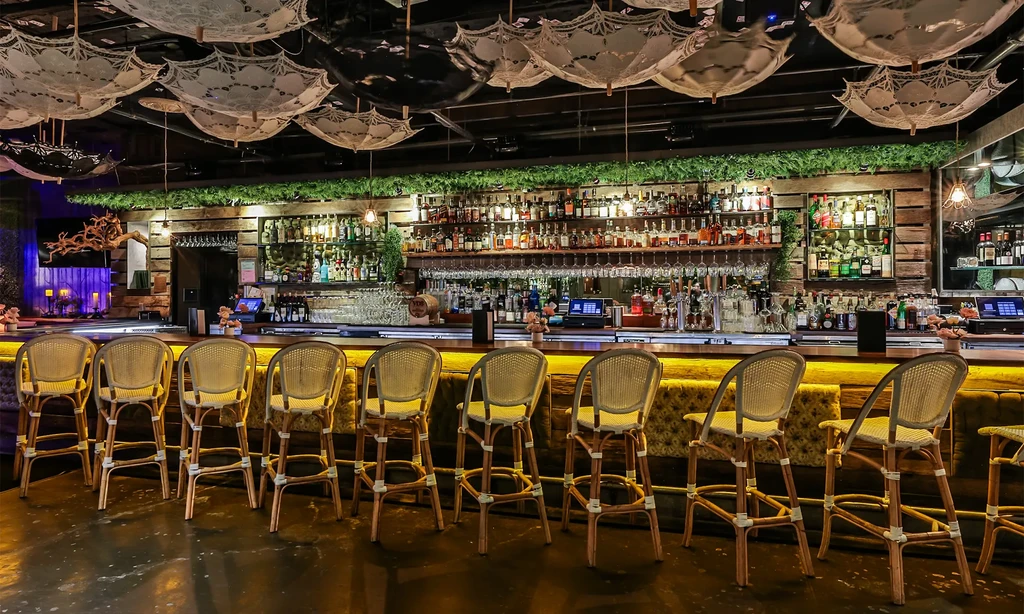 Well-stocked bar counter, empty rattan stools, lace parasols ceiling.