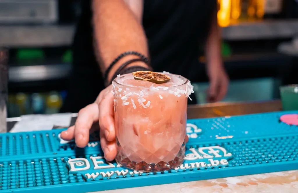 Bartender's hands present vibrant pink-orange cocktail with lime, flaked rim.