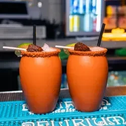 Two specialty drinks in unique cups with seasoned rims and garnishes.