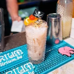Garnished milky cocktail with skull flag on bar mat beside shakers.