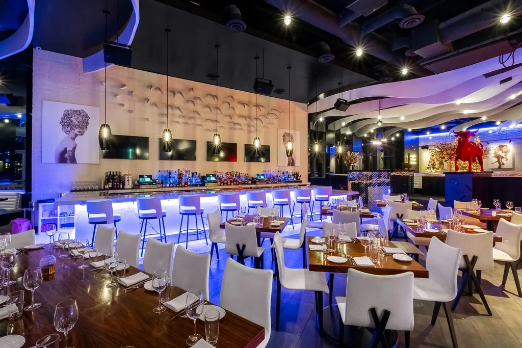 Empty modern restaurant interior with blue-lit bar, tables, and red bull statue.