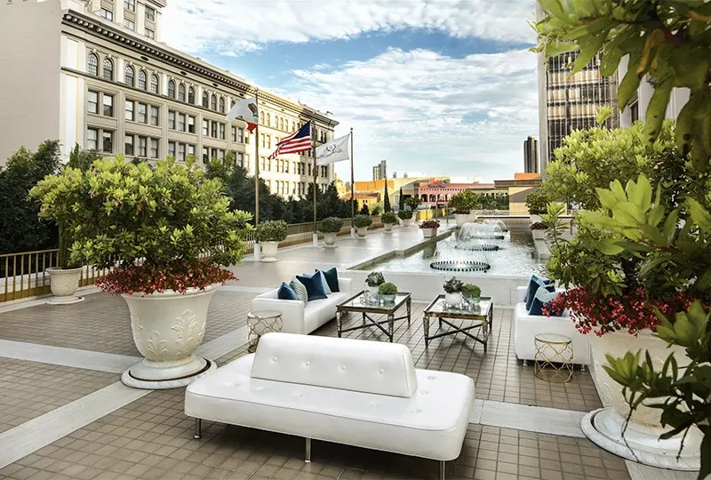 Rooftop terrace with modern white furniture, fountains, plants, and city flags.
