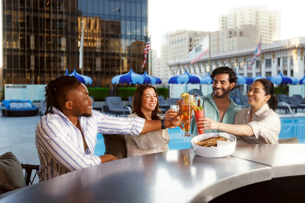 Four diverse people toast cocktails at an upscale rooftop bar with a pool.