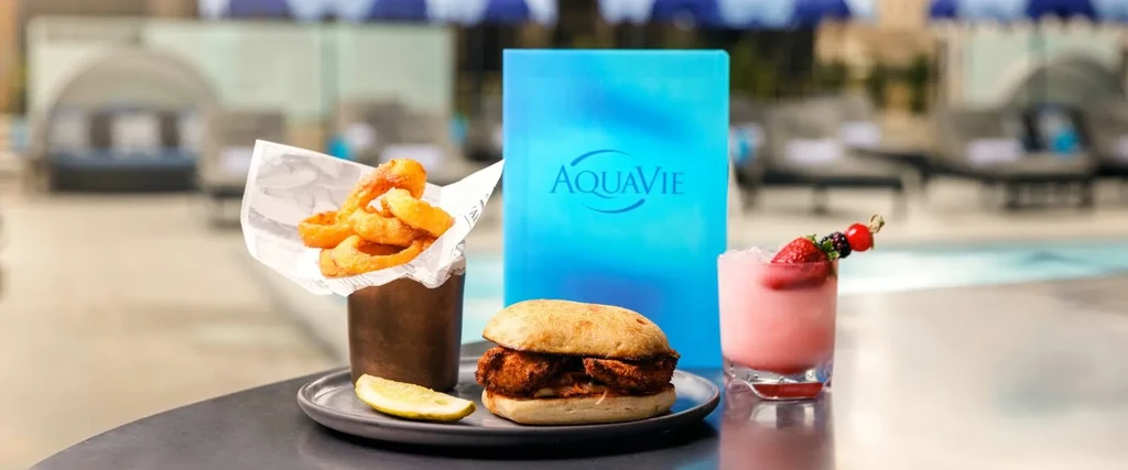 Onion rings, sandwich, pink cocktail, and Aquavie menu by a pool.