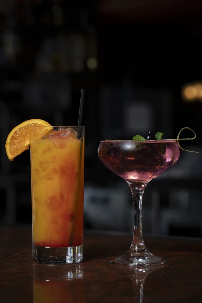 Two colorful cocktails, orange and pink, garnished on a dark bar.