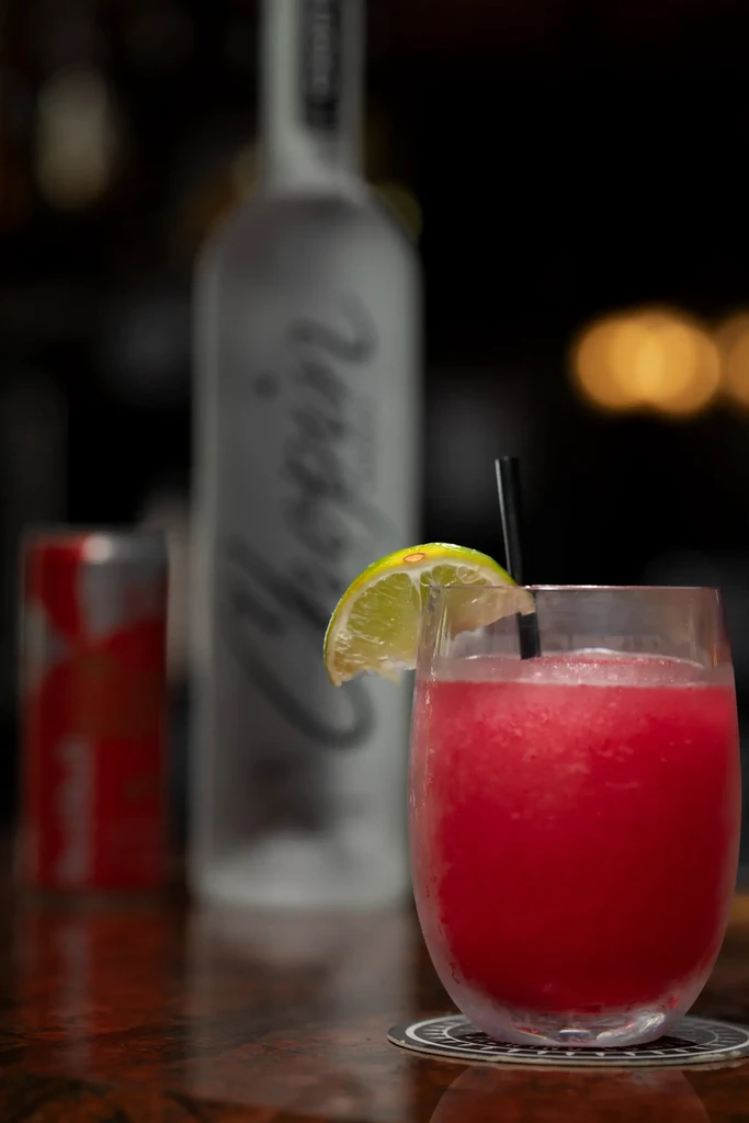 Vibrant red frozen cocktail with lime on wood; blurred bottle, can.