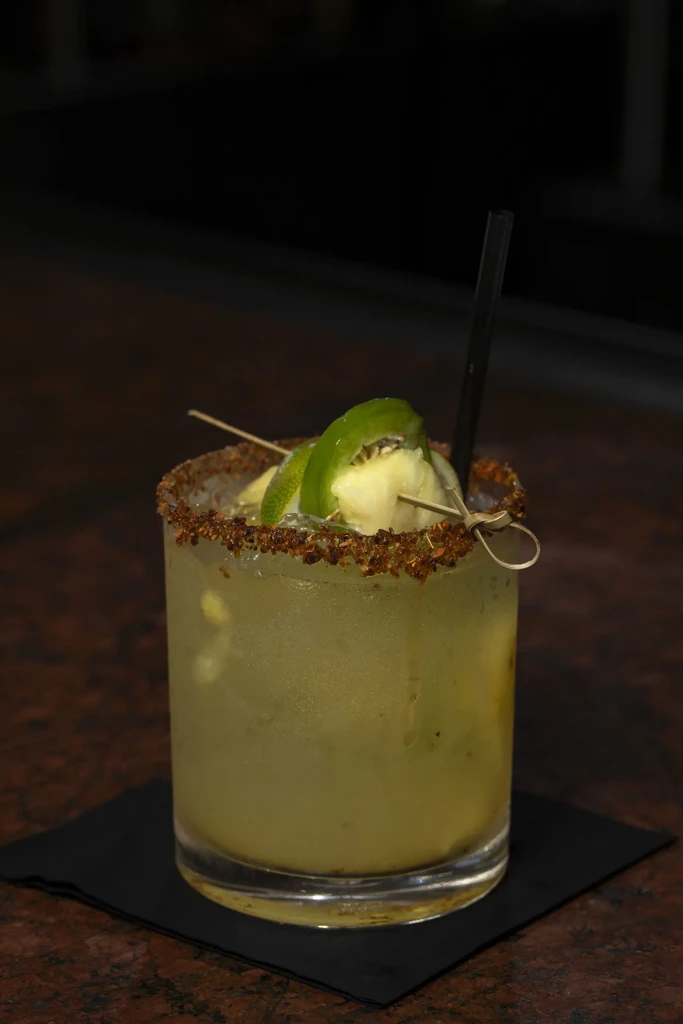 Yellow-green cocktail with seasoned rim, pineapple, and jalapeño garnish.