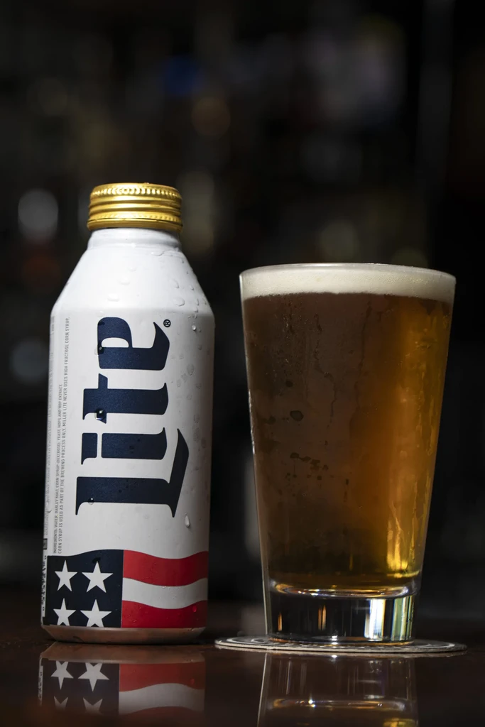 White Miller Lite can and condensation-covered beer glass on reflective surface.
