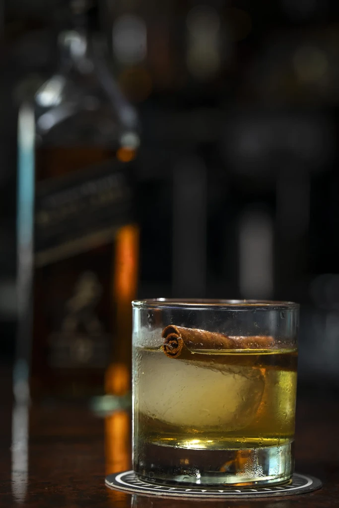 Close-up amber cocktail with spherical ice, cinnamon stick, blurred background.