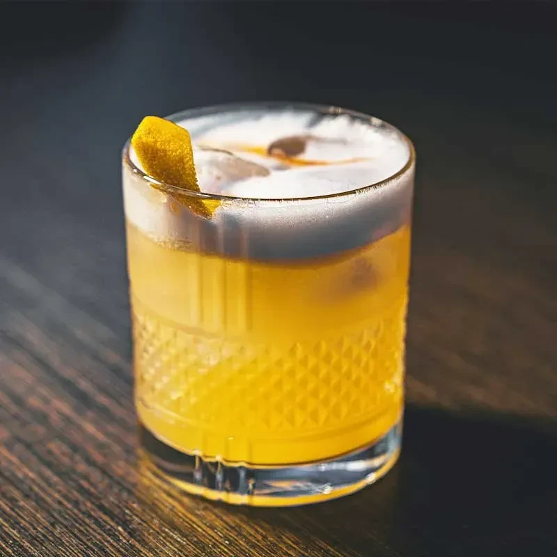 Yellow-orange cocktail with foam, citrus peel, and bitters in a faceted glass.