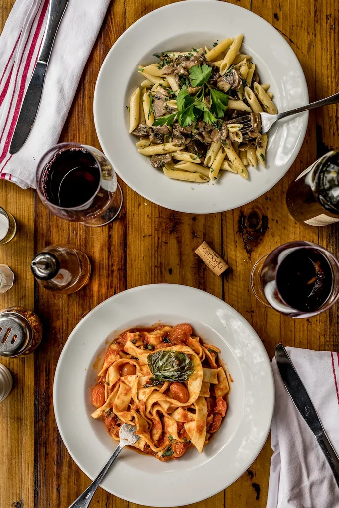 Two different pasta dishes and red wine on a rustic table.