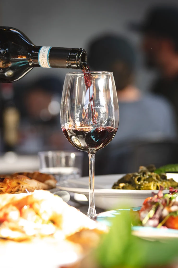 Red wine pours from bottle into glass in a busy restaurant.