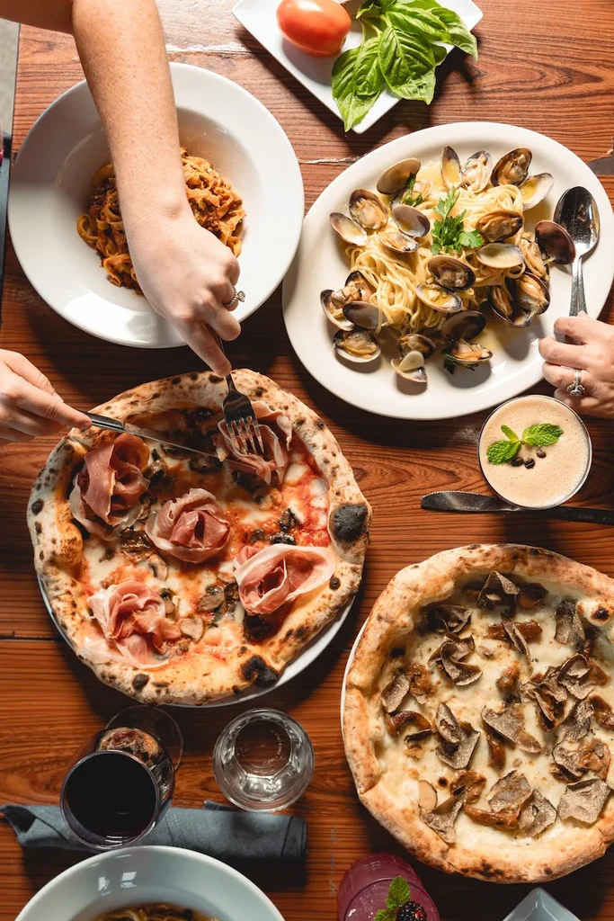Overhead shot: hands sharing a vibrant Italian meal of pasta, pizzas, and drinks.