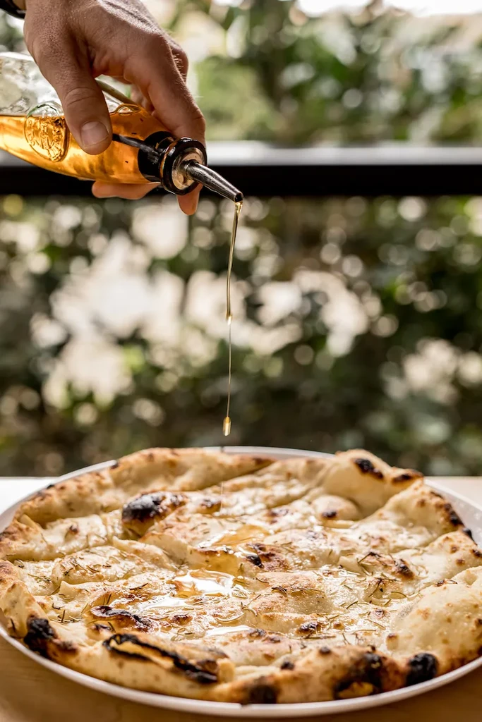 A hand drizzles golden oil over freshly baked herb focaccia.